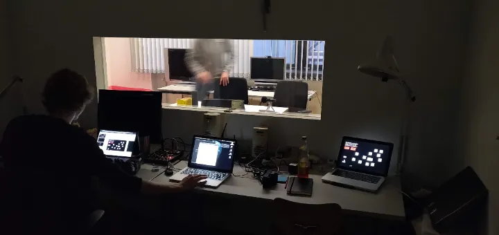 A person operates two laptops in a dimly lit room, while another person is visible through a window working in a well-lit adjacent room.