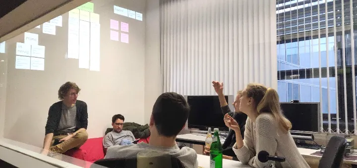 A group of four people having a discussion in an office room with a glass wall. Sticky notes and a projection are visible on a whiteboard. One person gestures while others listen.